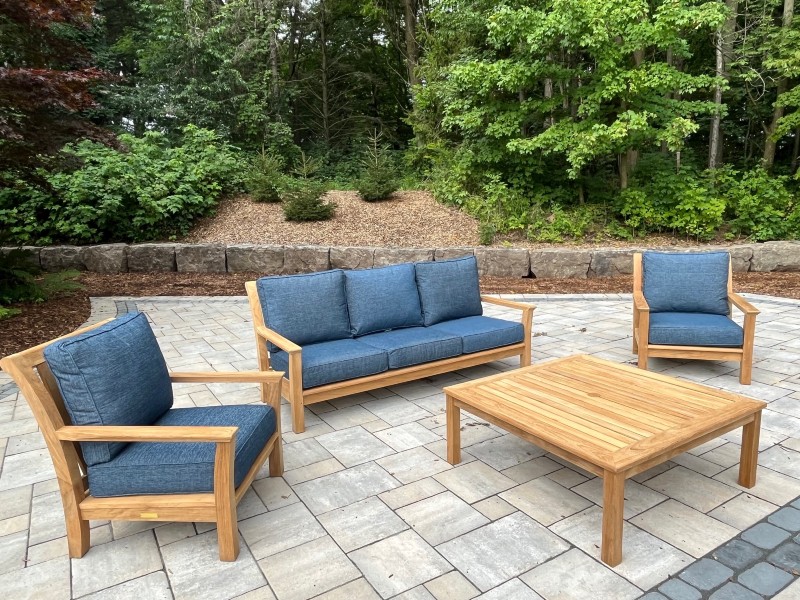 Modern square arm and back sofa and chairs with denim cushions and a square coffee table on a stone patio in front of a forest