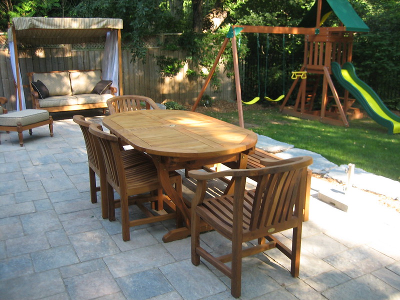 Oval teak dining table surrounded by armless and arm chairs with gently curved backs and teak love seat with beige cushions